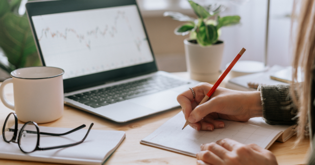 A woman's hand is writing in a notebook with a computer screen open to a graph in the backgroun.