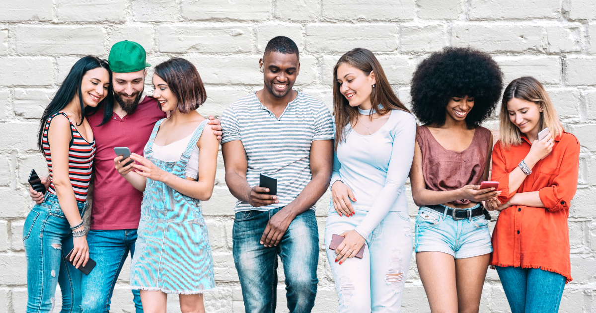 Seven people leaning on a brick wall looking at their phones.